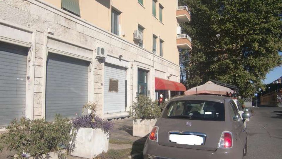 Strada urbana con edificio residenziale color beige, piano terra rivestito in pietra e serrande metalliche chiuse, con balconi ai piani superiori. Sul marciapiede ci sono grandi fioriere, un’auto parcheggiata e un grande albero accanto alla carreggiata.