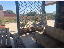 Terrazza sul tetto con pavimento piastrellato e area relax sotto una struttura in legno con grigliato, amaca a poltrona sospesa, divanetto e tavolino. Lungo il bordo ci sono fioriere e vasi con piante, con vista su edifici bassi e alberi sotto cielo sereno.