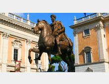Statua equestre in bronzo al centro di una piazza, con cavallo in passo e figura seduta, davanti a edifici storici con facciate ocra e bianche, cornici decorative e balaustre sul tetto. Sullo sfondo si vedono finestre alte, statue sul cornicione e alcuni alberi sotto un cielo azzurro.