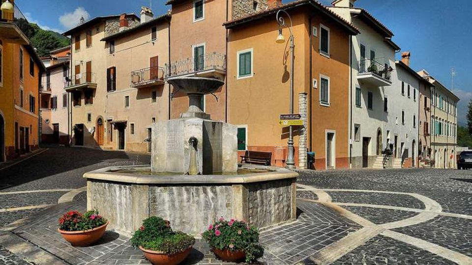 Piazza acciottolata di un borgo con una fontana in pietra al centro, circondata da vasi di fiori e panchine. Sullo sfondo si affacciano case a più piani dai colori pastello con balconi e persiane, illuminate dal sole.