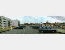 Ampio parcheggio sopraelevato con pavimentazione in cemento e auto parcheggiate in file, affiancato da una ringhiera gialla e un muretto. Sullo sfondo si vede un panorama urbano con edifici residenziali e strutture storiche a tetto basso sotto un cielo parzialmente nuvoloso.