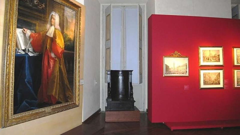 Interno di una sala museale con pavimento in parquet scuro e illuminazione a binario: a sinistra un grande dipinto incorniciato, a destra una parete espositiva rossa con quadri più piccoli. Sul fondo si vede una nicchia con una finestra alta e un basamento scuro.