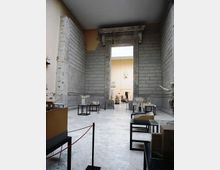 Interno di un museo con ampie pareti in pietra e pavimento chiaro, organizzato come galleria con modelli architettonici su tavoli e vetrine. Al centro si vede un grande portale monumentale in pietra decorata che conduce a una sala successiva.