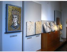 Sala museale con pareti bianche che espone un grande mosaico a tessere dorate con ritratto frontale e, accanto, rilievi in pietra appesi e busti marmorei allineati su un lungo basamento in legno. Illuminazione puntuale sulle opere e pavimento in legno.