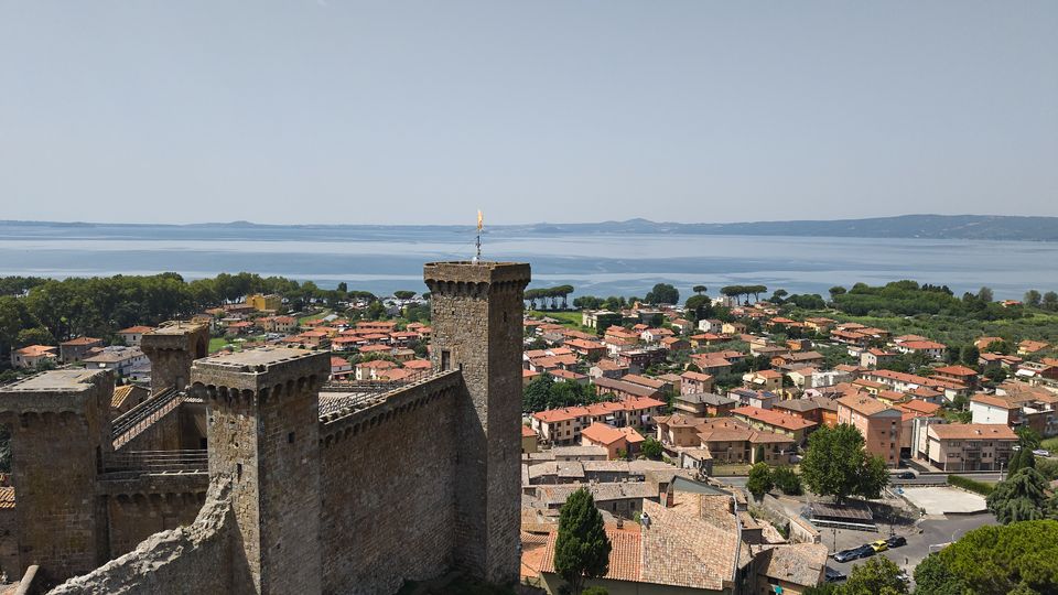 Veduta dall’alto di una fortezza medievale in pietra con mura merlate e torri quadrate, affacciata su un centro abitato con case dai tetti in terracotta. Sullo sfondo si vede un ampio lago con costa alberata e colline all’orizzonte sotto un cielo sereno.
