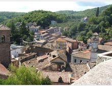 Veduta panoramica di un borgo collinare con case in pietra e tetti in coppi, stradine strette e un campanile con orologio. Il paese è circondato da colline boscose e vegetazione fitta.