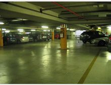 Parcheggio sotterraneo con pavimento in cemento lucido, colonne arancioni e illuminazione al neon sul soffitto con tubazioni a vista. Auto parcheggiate lungo i lati e corsia centrale libera delimitata da linee gialle.