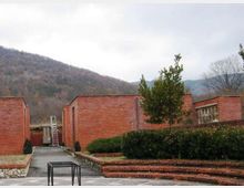 Cortile esterno con vialetto in cemento tra edifici bassi in mattoni rossi, con siepi e gradoni in primo piano. Sullo sfondo si vedono colline boscose sotto un cielo nuvoloso.