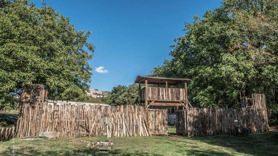 Recinto in legno con pali verticali e un portale centrale sormontato da una piccola torretta coperta, in un’area erbosa. Grandi alberi circondano la struttura e sullo sfondo si vede una collina sotto un cielo sereno.