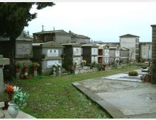 Cimitero con file di loculi e cappelle funerarie in muratura, con lapidi e vasi di fiori. Un vialetto pavimentato corre accanto a un’area erbosa, con alberi sullo sfondo.