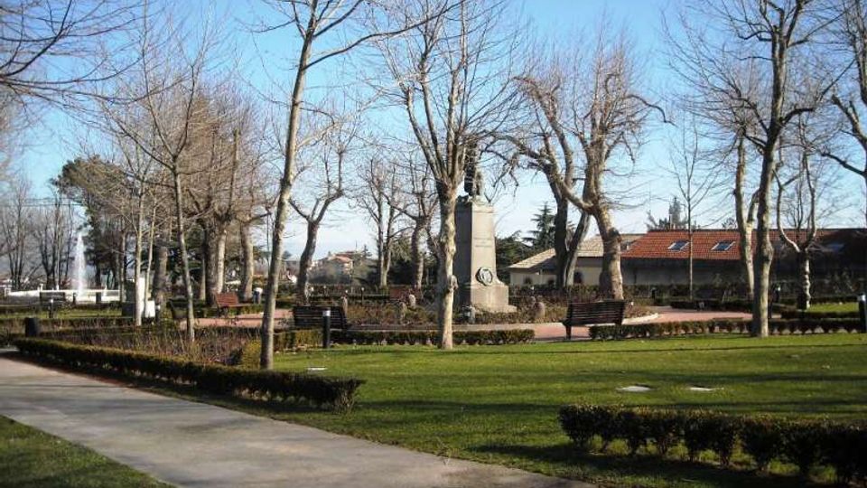 Parco urbano con prato curato, vialetti in cemento e siepi basse, circondato da alberi spogli. Al centro si vede un monumento su piedistallo con panchine vicine, con una fontana e edifici dai tetti rossi sullo sfondo.