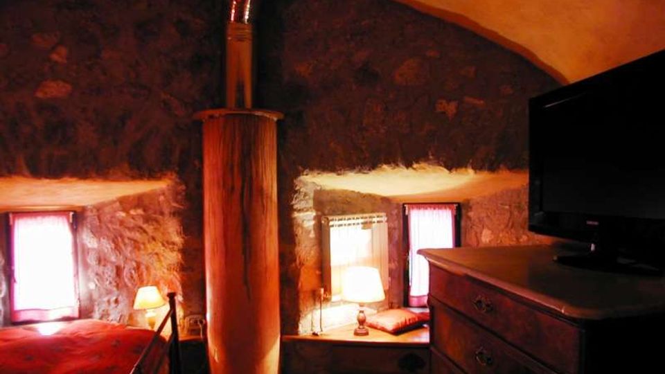 Interno di una camera rustica con soffitto a volta e pareti in pietra, con un grande camino centrale e pavimento a piastrelle. Due piccole finestre con tende, un letto, un comò in legno con televisore e lampade da comodino illuminano l’ambiente.
