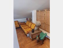 Salottino in mansarda con soffitto inclinato e pavimento in parquet, arredato con divani a fiori arancioni, tavolino basso e libreria. Lungo la parete c’è un paravento in bamboo e un mobiletto con lampada decorativa accanto a uno sgabello verde.