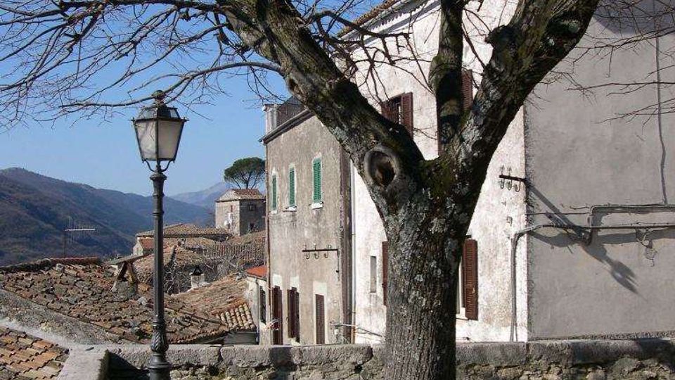 Terrazza panoramica in un borgo collinare con panchina, grande albero spoglio e lampione in ferro accanto a un muretto in pietra. Sullo sfondo si vedono case in muratura con persiane e tetti in coppi, con montagne all’orizzonte sotto cielo sereno.