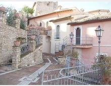 Cortile in pietra di un edificio storico con facciata rosa, scale e terrazze con ringhiere in ferro battuto e vasi in terracotta lungo i muri. Pavimentazione a mosaico e lampioni in stile classico, con strutture in pietra e tetti in tegole sullo sfondo.