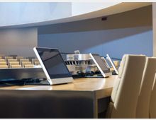 Sala conferenze moderna con file di sedute sullo sfondo e un grande tavolo in legno in primo piano con schermi/monitor inclinati e cablaggi. Pareti chiare e soffitto curvo con illuminazione integrata definiscono un ambiente istituzionale e ordinato.
