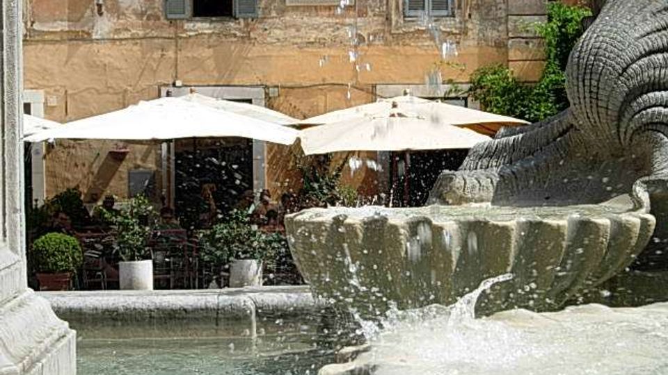 Fontana in pietra in una piazza, con vasca d’acqua in primo piano e schizzi visibili. Sullo sfondo, un edificio storico con finestre e tende da esterno bianche sopra tavoli di un’area caffè.