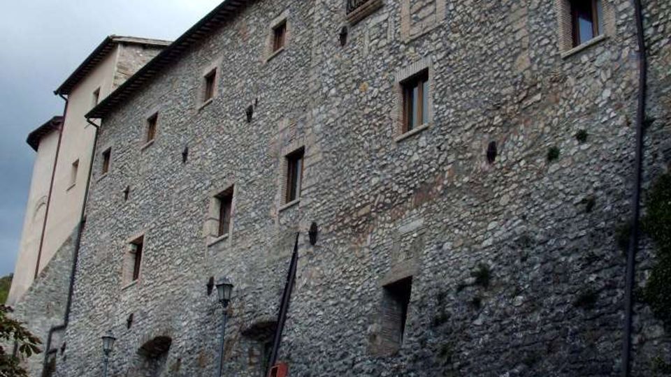 Grande edificio storico in pietra su un pendio, con facciata lunga e irregolare, piccole finestre e balconcini in ferro battuto. In primo piano si vede un muro di contenimento in pietra lungo una strada con lampioni, sotto un cielo nuvoloso.