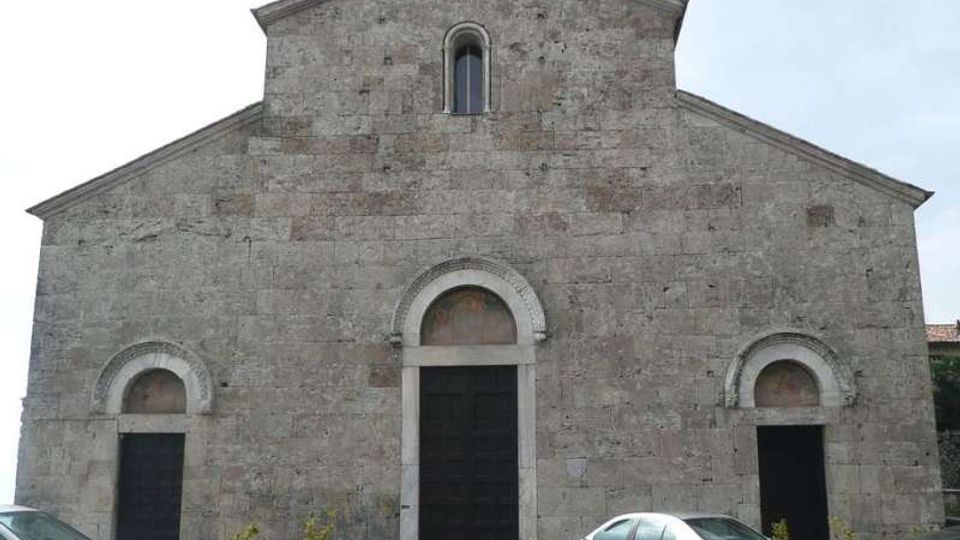 Facciata in pietra di una chiesa in stile romanico, con frontone triangolare sormontato da una croce, una piccola finestra ad arco e tre ingressi con portali ad arco. Davanti si vede una piazza pavimentata con alcune auto parcheggiate e grandi fioriere.