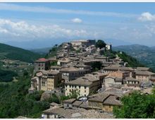 Veduta panoramica di un borgo su una collina, con case addossate tra loro e tetti in tegole. Sullo sfondo si vedono colline e montagne sotto un cielo azzurro con nuvole sparse.