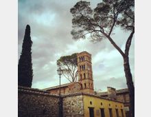 Complesso storico in muratura con edificio basso intonacato giallo e tetti in coppi, affiancato da un campanile romanico in mattoni con archi sovrapposti. In primo piano si vede un muro in pietra e un lampione, con alti pini e un cipresso sullo sfondo sotto un cielo nuvoloso.