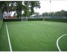 Campo da calcetto in erba sintetica con linee bianche e piccola porta sul fondo, circondato da recinzioni e pannelli verdi. Sullo sfondo si vedono alberi alti e pali dell’illuminazione lungo il perimetro.