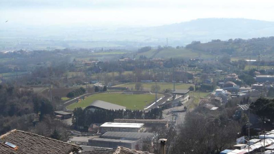 Vista panoramica dall’alto su una valle collinare con campi e piccoli insediamenti; al centro si vede uno stadio o campo sportivo ovale con tribune e impianti. In primo piano compaiono tetti in tegole, mentre sullo sfondo si estendono colline e montagne in lontananza.