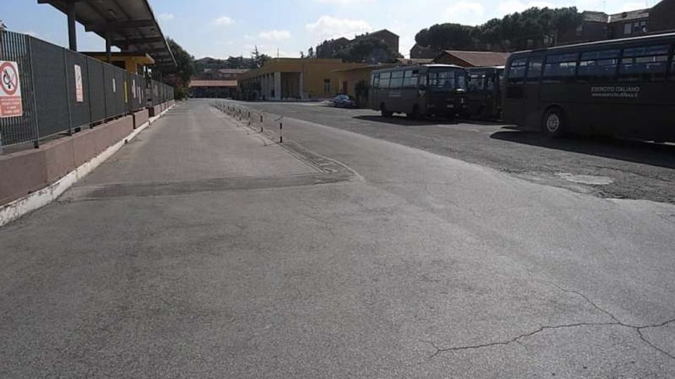 Ampio piazzale asfaltato di un deposito autobus, con pensiline e recinzioni sul lato sinistro e diversi autobus parcheggiati sulla destra. Sullo sfondo si vedono edifici bassi e alberi sotto un cielo parzialmente nuvoloso.