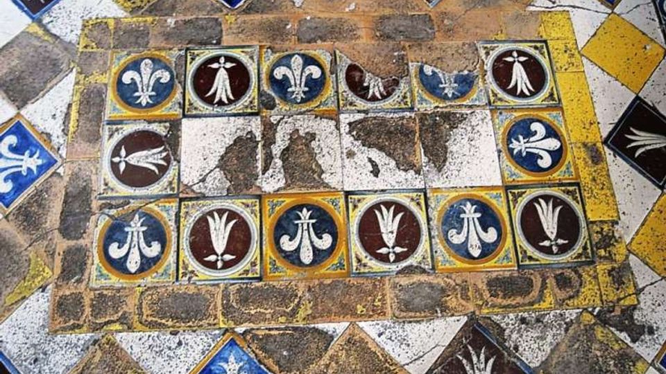 Pavimento decorato a mosaico con un riquadro centrale bordato da piastrelle colorate raffiguranti gigli stilizzati in blu, bianco e marrone. Intorno si vede una cornice di tessere romboidali e rettangolari in giallo, blu, nero e bianco, con segni di usura e lacune.