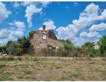 Edificio rurale in rovina con mura scrostate e tetto parzialmente crollato, con un piccolo campaniletto sulla sommità. Davanti c’è una recinzione metallica e vegetazione incolta, con alberi e alcune case sullo sfondo sotto un cielo azzurro con nuvole.