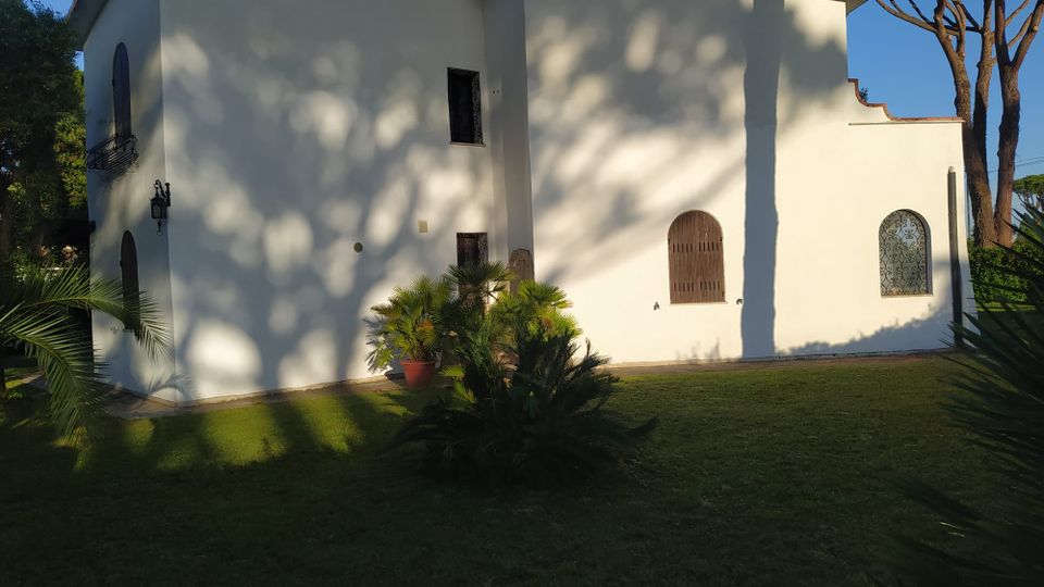 Edificio intonacato di bianco con tetto in tegole, finestre ad arco con grate in ferro e una piccola balconata laterale. Davanti si trova un prato con piante ornamentali e palme, con alte conifere sullo sfondo e ombre degli alberi proiettate sulla facciata.