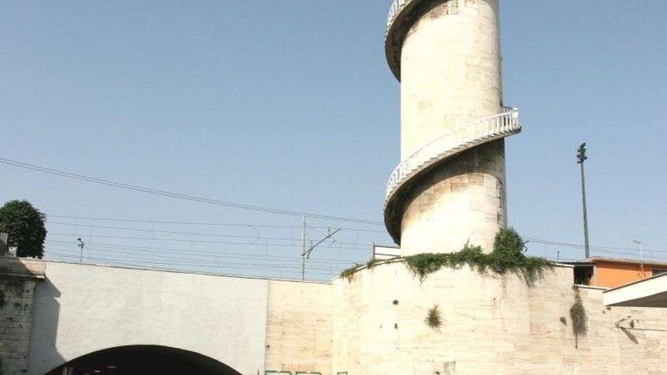 Torre cilindrica in cemento con scala a chiocciola esterna, accanto a un sottopasso stradale sotto un viadotto. L’area circostante è urbana e industriale, con muri in cemento con graffiti, parcheggio di auto e linee ferroviarie sopraelevate.