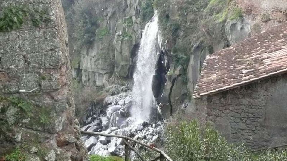 Cascata naturale che scende da una parete rocciosa in una gola, con massi alla base e vegetazione sulle scarpate. In primo piano si vedono un sentiero erboso con staccionata in legno e muri in pietra, con un edificio in pietra dal tetto in tegole sulla destra.