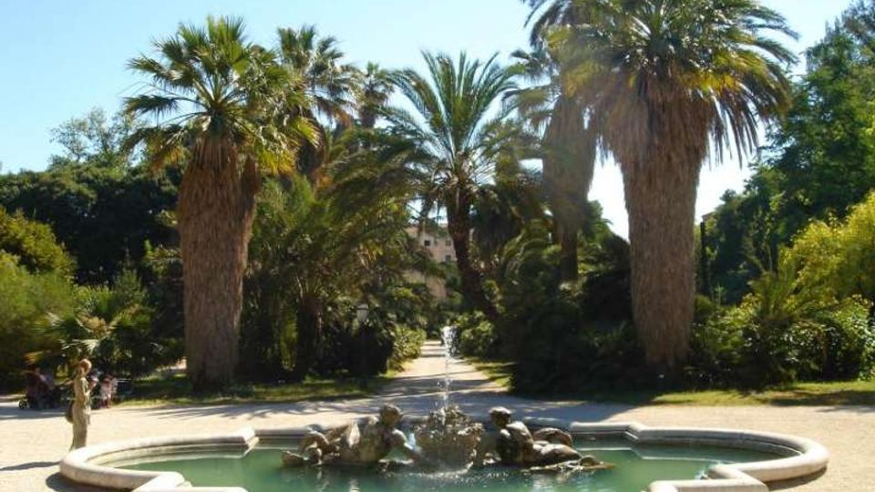 Fontana circolare in pietra con acqua verde e piccole statue, situata al centro di un viale in un parco. Intorno si vedono alte palme e vegetazione mediterranea, con sentieri ghiaiosi e cielo sereno sullo sfondo.
