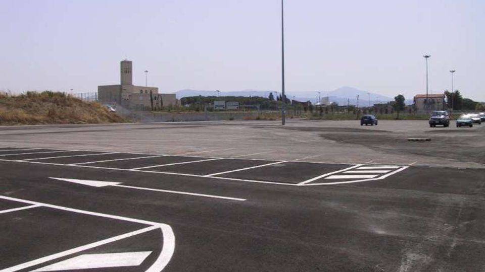 Grande parcheggio asfaltato quasi vuoto con segnaletica orizzontale bianca e alti lampioni al centro dell’area. Sullo sfondo si vedono edifici bassi e un paesaggio pianeggiante con colline lontane sotto un cielo chiaro.