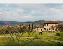 Casale rurale in pietra con tetto in tegole e archi al piano terra, affacciato su un ampio prato attraversato da un sentiero. Intorno ci sono alberi spogli o in fiore e colline ondulate sullo sfondo sotto un cielo parzialmente nuvoloso.