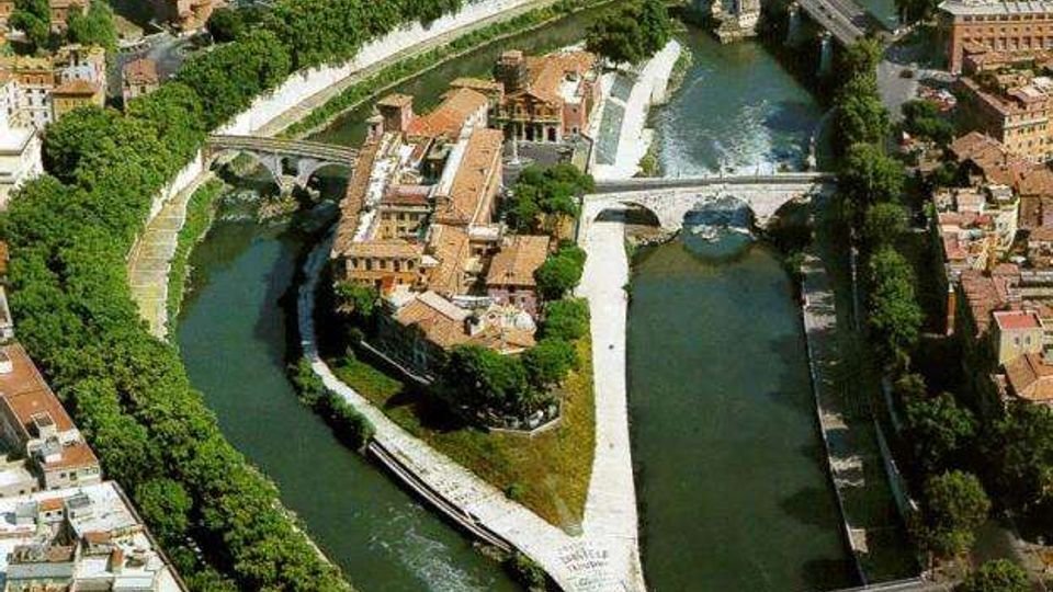 Veduta aerea di un’isola urbana triangolare circondata da un fiume, con edifici storici dai tetti in terracotta e argini in pietra. Due ponti attraversano il corso d’acqua e una piccola cascata è visibile vicino a uno degli sbarramenti, con alberi e strade che costeggiano le rive.