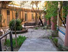 Cortile interno di un edificio con pavimentazione in ghiaia e un breve tratto di piastrelle, aiuole e alberi; al centro si vedono panchine e una grande fioriera circolare. Sui lati ci sono facciate con porte e finestre e un porticato con colonne cilindriche rosa.
