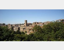 Vista panoramica di un borgo collinare con case in pietra dai tetti in coppi e una torre campanaria quadrata che svetta al centro. In primo piano ci sono alberi e vegetazione fitta, con cielo limpido sullo sfondo.