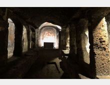 Interno di una cripta o cappella sotterranea scavata nella roccia, con navata centrale e pilastri grezzi che sostengono una volta bassa. In fondo si vede un piccolo altare davanti a un’abside con affresco, illuminata da una luce proveniente dall’alto.