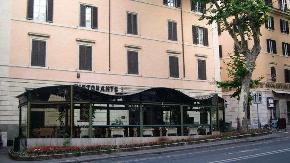 Facciata di un edificio residenziale di più piani color pesca con persiane scure, affacciato su una strada urbana. Al piano terra c’è un ristorante con veranda coperta da una struttura metallica e aiuola lungo il marciapiede, con un grande albero a lato.