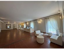 Ampia sala interna con pavimento in piastrelle color terracotta, pareti chiare e illuminazione a binario. Lungo il lato destro ci sono grandi finestre con tende bianche e un’area lounge con sedute imbottite chiare e tavolini, mentre sul fondo si vedono specchi a parete e un bancone tipo buffet.