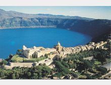 Veduta panoramica di un borgo affacciato su un ampio lago circondato da colline, con un grande complesso storico in primo piano caratterizzato da cupola e mura. Case in pietra e vegetazione terrazzata degradano verso la riva lungo una strada costiera.