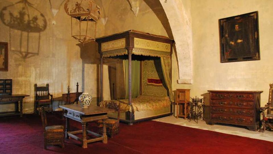 Interno di una stanza storica con soffitto a volte e pareti in pietra intonacata, arredata con un grande letto a baldacchino, tavolo e sedie in legno. Nella sala si vedono anche un cassettone, un ritratto appeso alla parete e una lanterna sospesa al soffitto su pavimento rivestito da tappeto rosso.