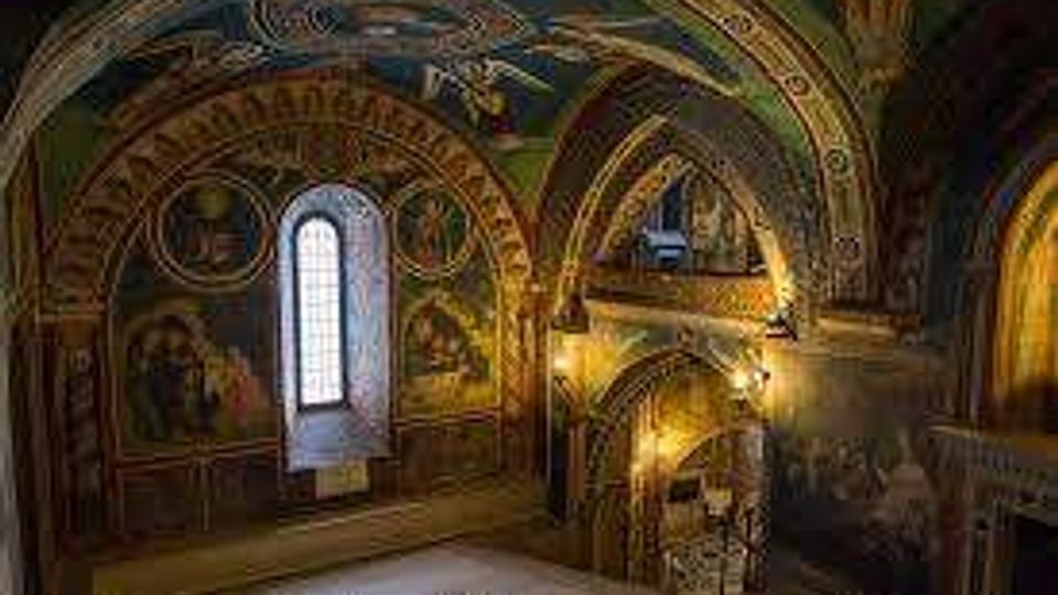 Interno di una chiesa o cappella storica con volta a botte e archi decorati da affreschi, una stretta finestra ad arco e un piccolo soppalco con scala. Pareti dipinte con motivi religiosi e illuminazione calda che mette in risalto le decorazioni.