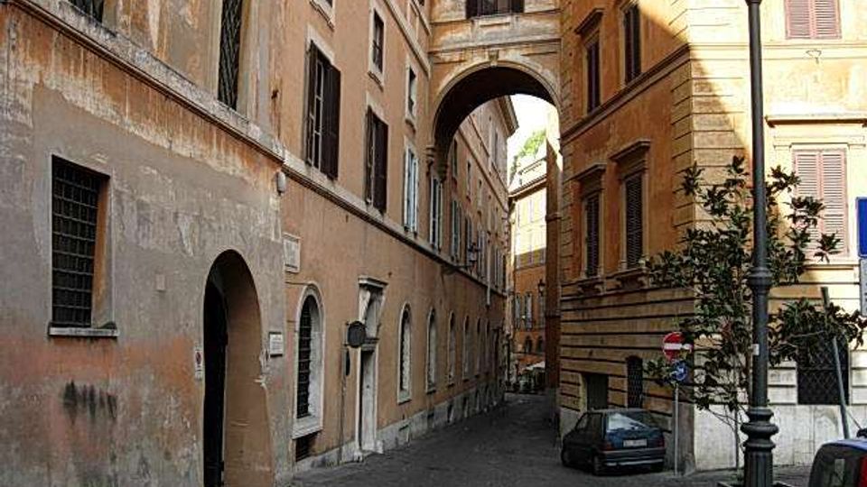 Stretta via urbana tra edifici storici in intonaco chiaro, con finestre a persiane e portoni ad arco al piano terra. In fondo si vede un grande arco che collega i palazzi sopra la strada, con un lampione e alcune auto parcheggiate lungo il bordo.