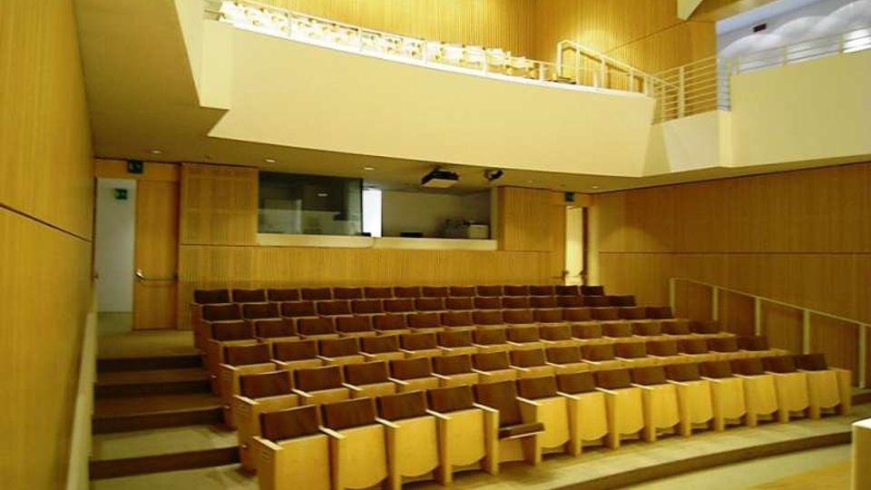 Interno di un auditorium moderno con pareti rivestite in legno chiaro e file di sedute in legno disposte su gradinate. In alto si vede un balcone con parapetto e una piccola cabina tecnica affacciata sulla sala, illuminata da faretti a soffitto.