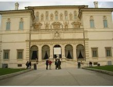 Facciata di un grande palazzo storico in stile rinascimentale, con decorazioni scolpite, finestre simmetriche e tre ampie arcate al piano terra. Una scalinata centrale conduce all’ingresso principale, affacciato su un viale pedonale con prati ai lati.