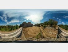 Vista panoramica di un’area all’aperto con un edificio in pietra di tipo fortificato e un ampio spiazzo sterrato centrale. Ai lati si vedono rampe pavimentate con cordoli in pietra e un alto muro in blocchi di pietra, con vegetazione e cielo parzialmente nuvoloso sullo sfondo.
