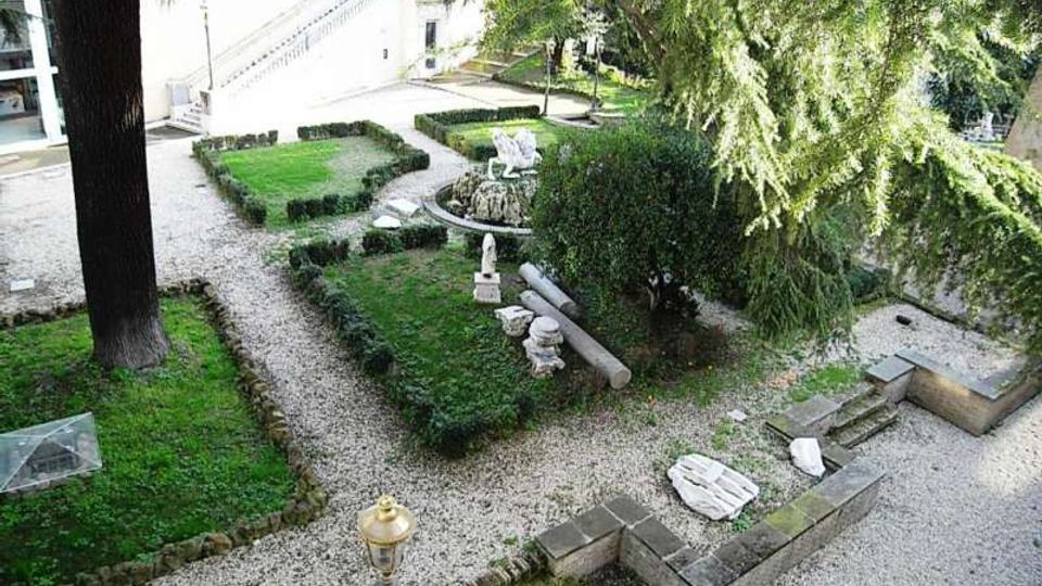 Giardino formale con vialetti in ghiaia e aiuole geometriche bordate da siepi basse, con una piccola fontana o vasca circolare al centro e sculture in pietra. Alberi alti e un edificio chiaro con scala esterna fanno da sfondo.